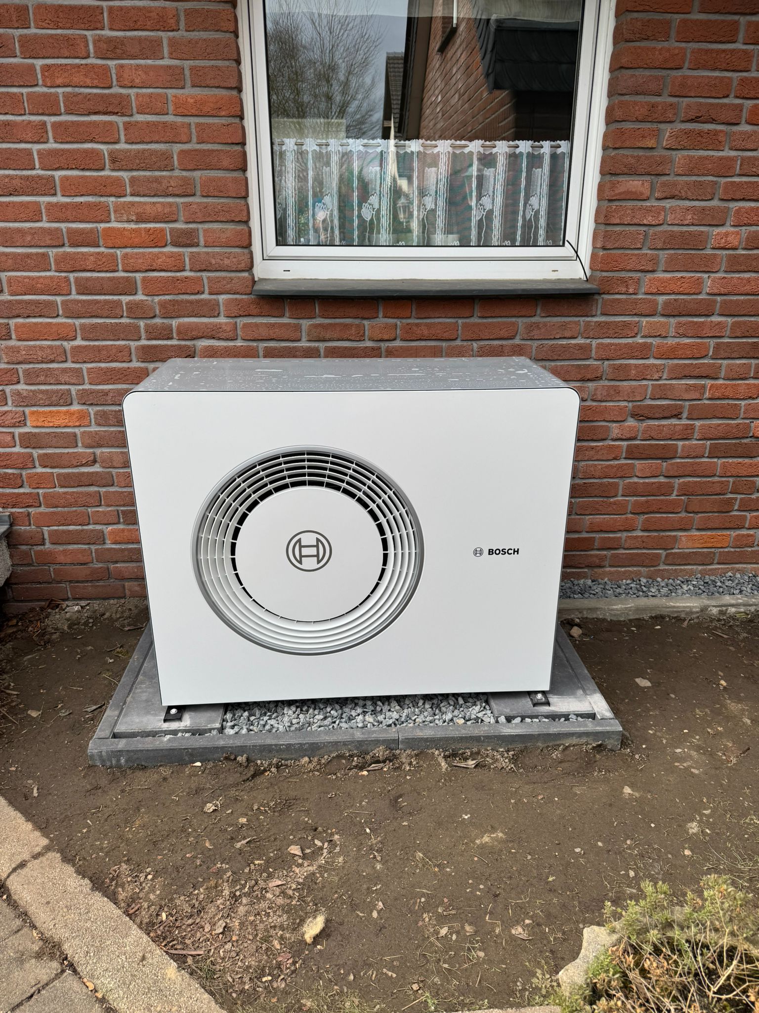 Eine Bosch-Wärmepumpe steht auf einer gepflasterten Fläche vor einer Hauswand aus rotem Klinker, direkt unter einem Fenster.